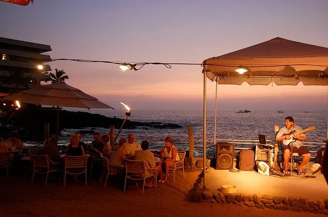 Guests dining outdoors under umbrellas at sunset while a musician plays guitar nearby