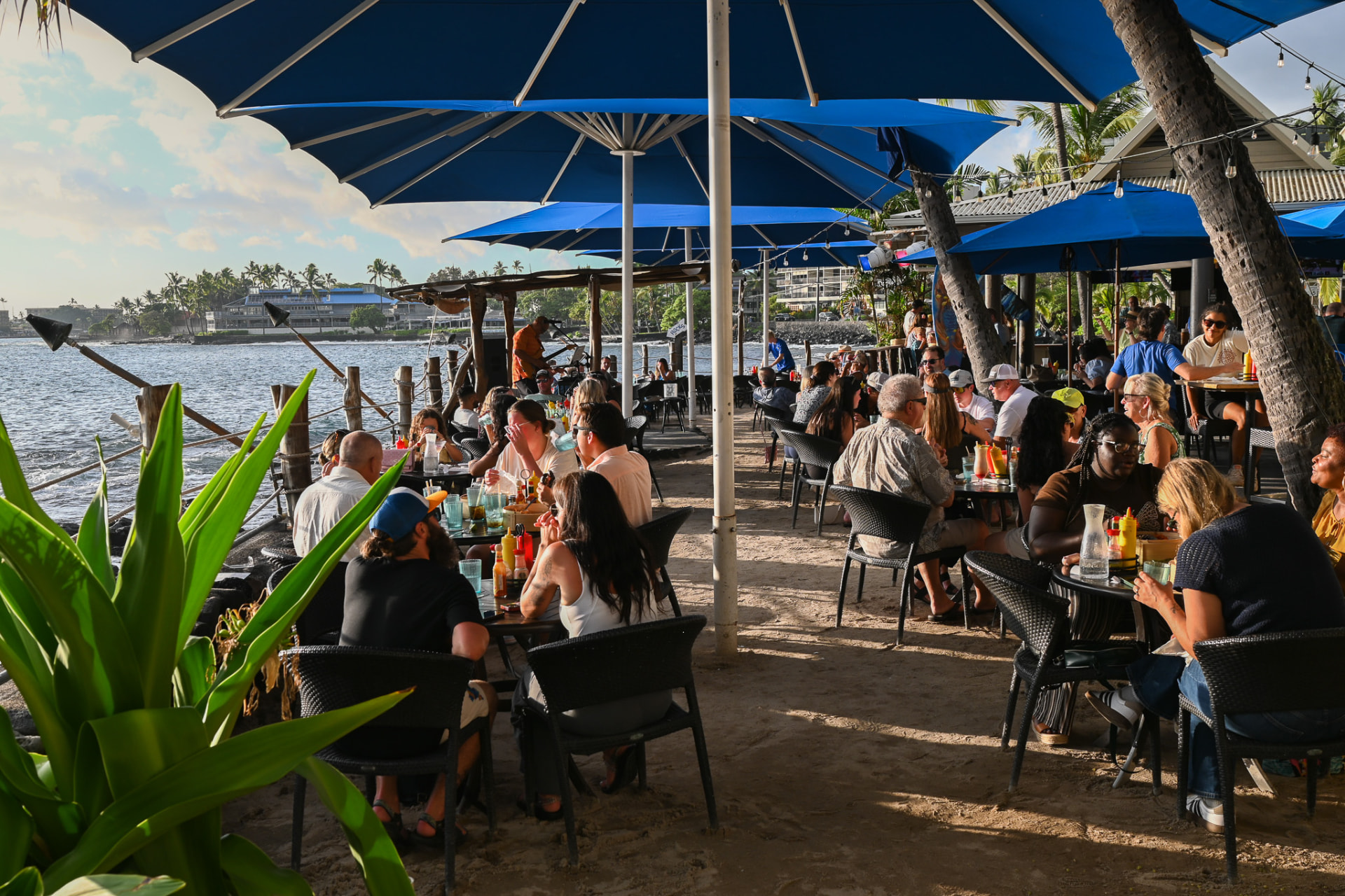 Guests at oceanfront tables under blue umbrellas on the sand